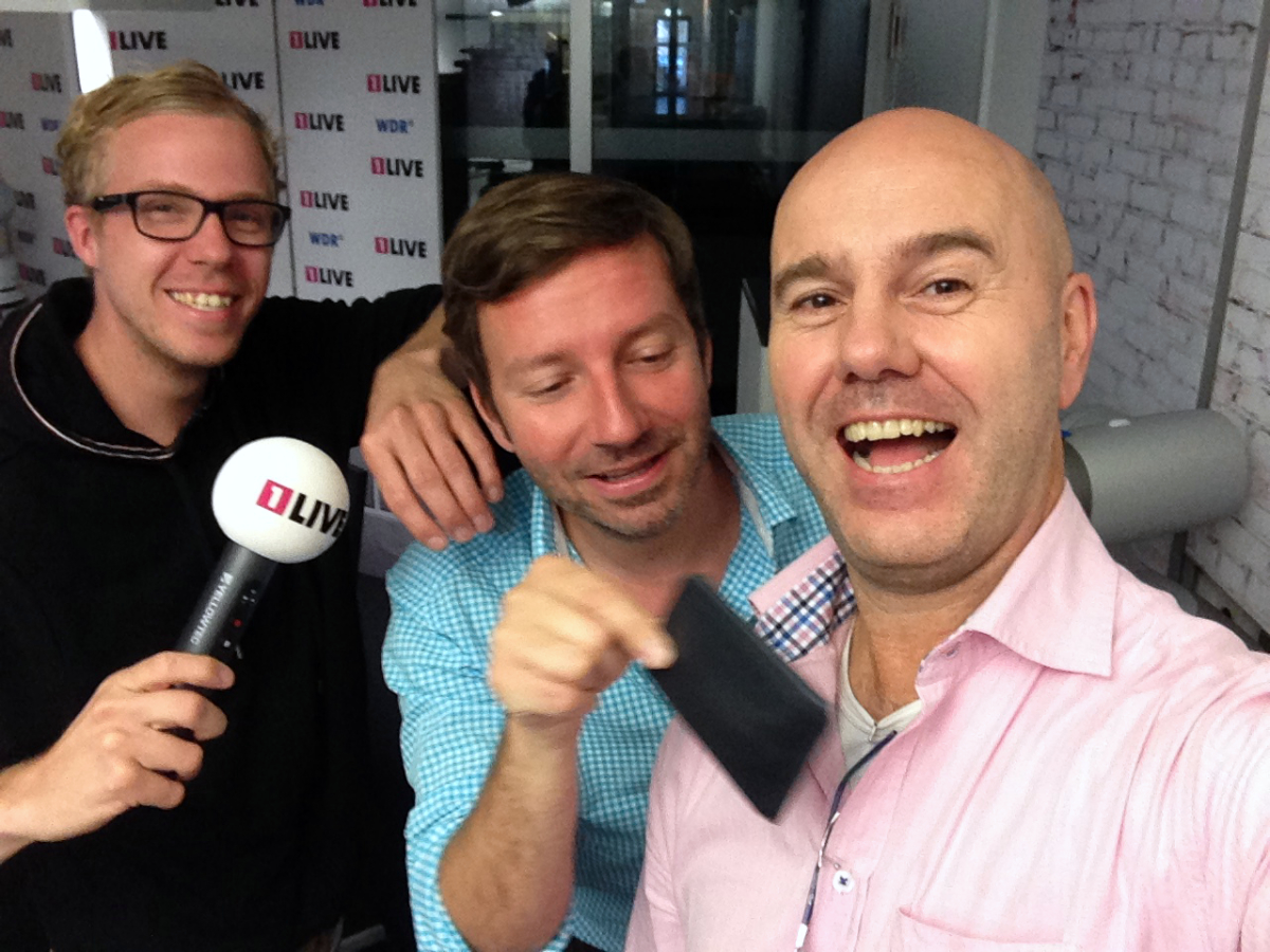 Zwei 1 Live Radiomoderatoren, Daniel Danger und Thorsten Schorneiner mit einem Mikrofon, bestehlen den lachenden Taschendieb Giovanni Alecci.